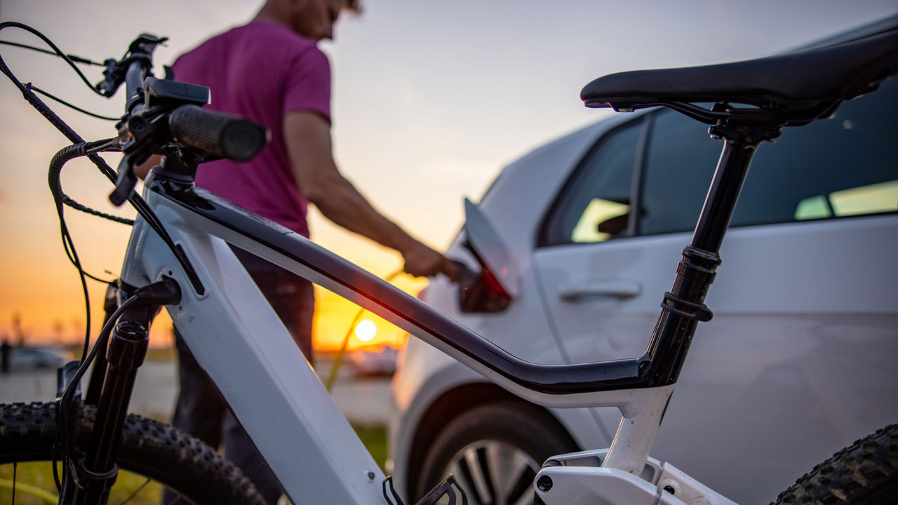 Im Vordergrund steht ein Fahrrad und im Hintergrund tankt ein Mann sein E-Auto