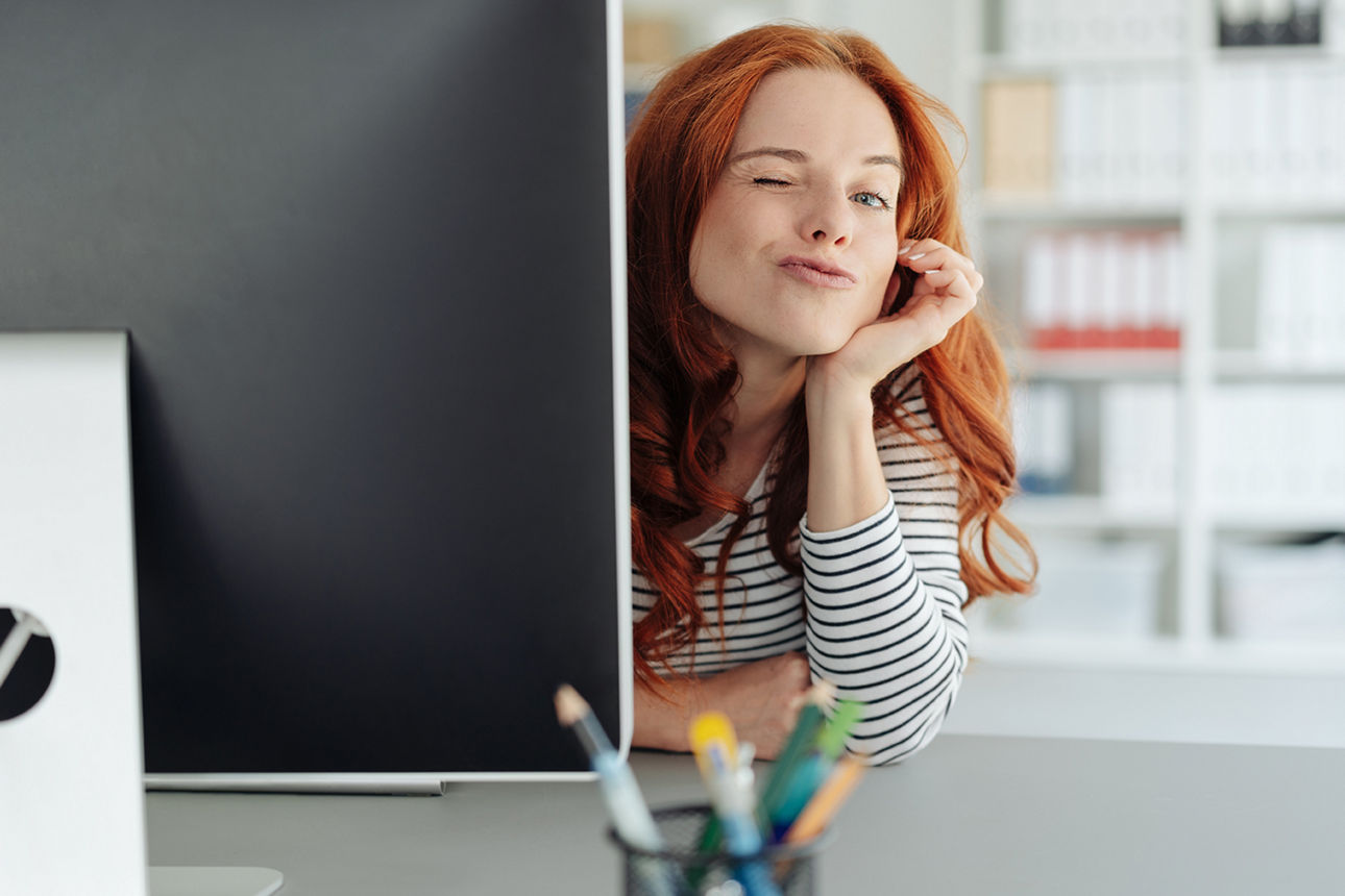 Frau die hinter ihrem Bildschirm hervorschaut und in die Kamera zwinkert.
