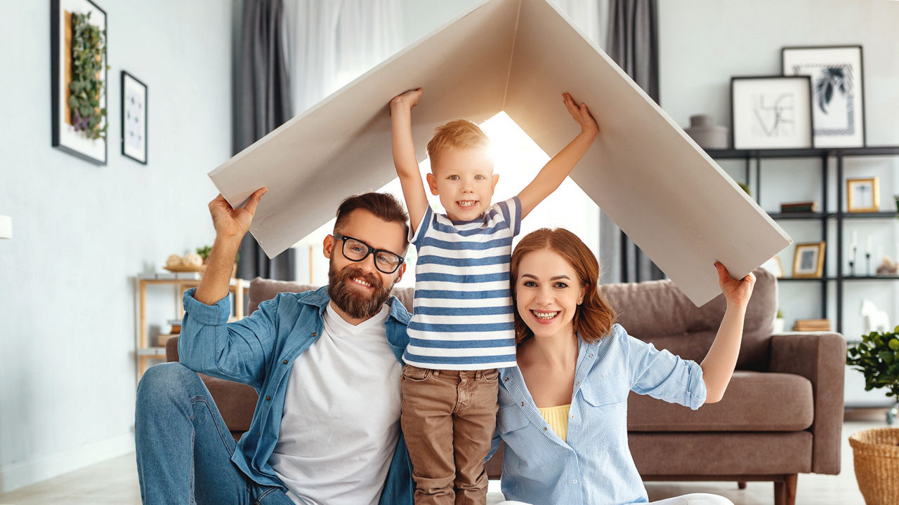 Eine junge Familie hält sich im Wohnzimmer ein Dach aus Pappe über den Kopf. 