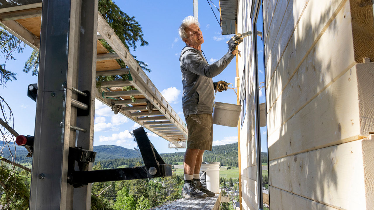 Mann streicht das Haus von außen und hält einen Pinsel sowie Eimer in den Händen.