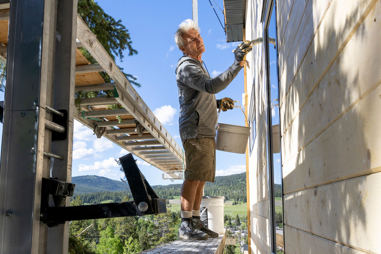 Mann streicht das Haus von außen und hält einen Pinsel sowie Eimer in den Händen.