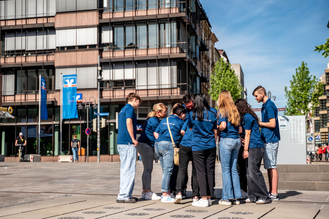Zu sehen ist eine Gruppe von Auszubildende auf dem Uniplatz in Fulda. Im Hintergrund ist die Bankfiliale in der Bahnhofstraße
