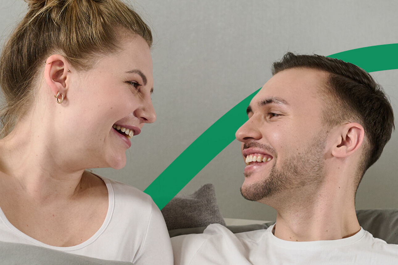 Ein Paar liegt gemeinsam im Bett und lächeln sich an. 