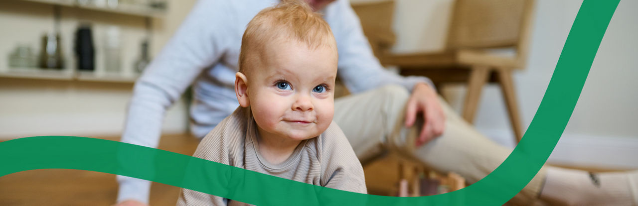 Ein Baby lächelt in die Kamera, im Hintergrund sitzt der Vater