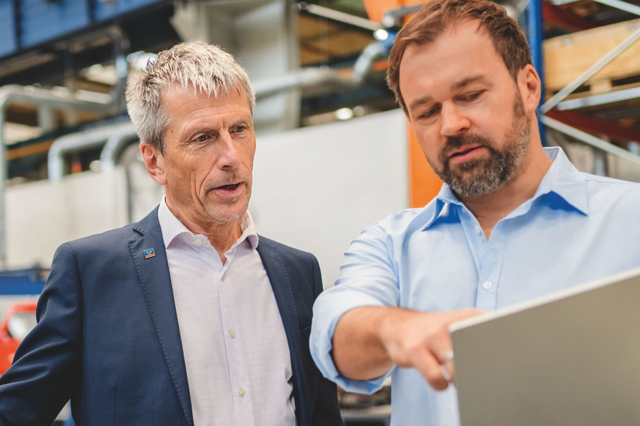 Zwei Personen stehen in einer Industriehalle zwischen Regalen und Maschinen und betrachten gemeinsam einen Laptop.