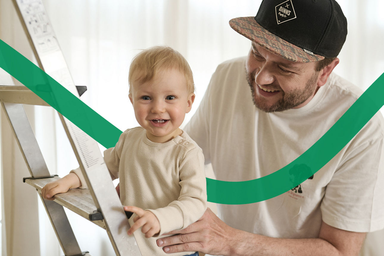 Während der Vater das Kind festhält, steht es auf der Leiter im Wohnzimmer. 