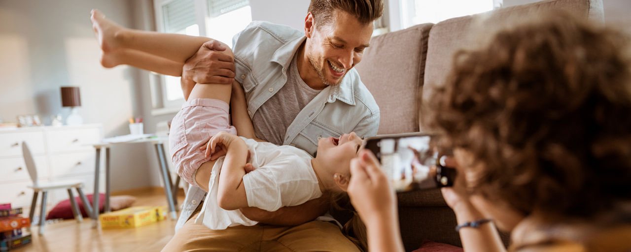 Vater und Kind albern im Wohnzimmer und werden dabei von dritter Person mit Handy fotografiert