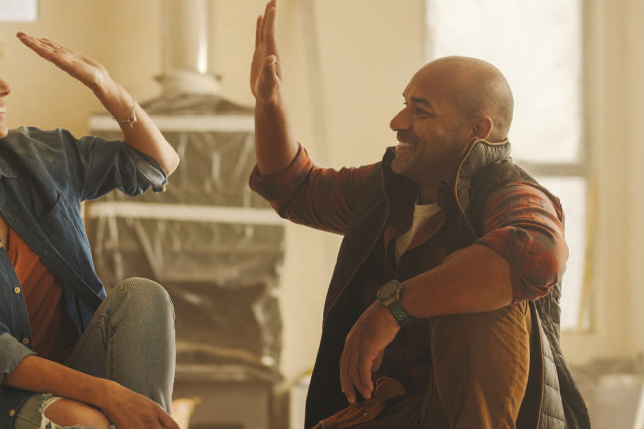 Ein Mann mit unglücklicher Mimik steht vor einem mit Klebeband zusammengehaltenen Haus, in den Händen Schere und Tape.