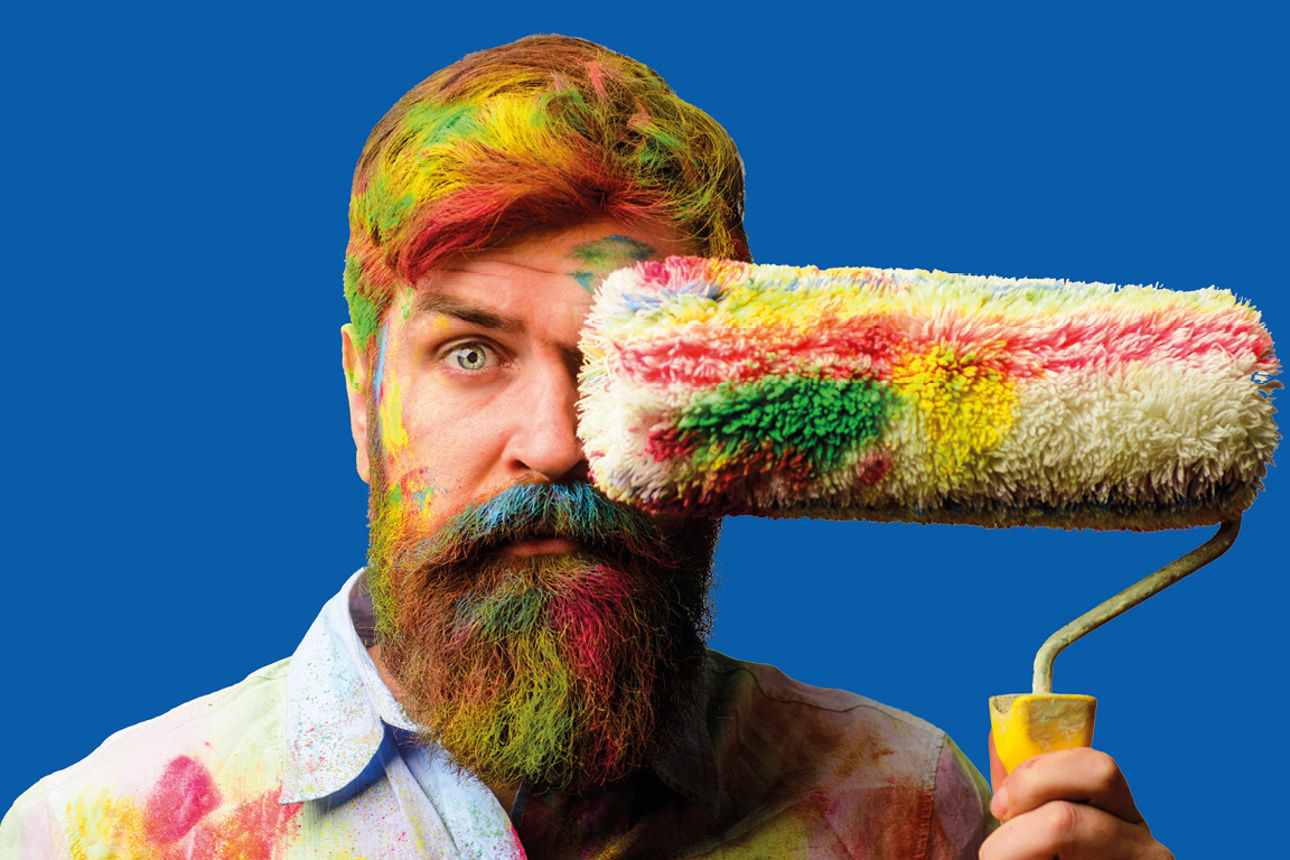 Mann steht vor einem blauen Hintergrund, hält eine Farbrolle in der Hand und ist bunt mit Farbe verschmiert