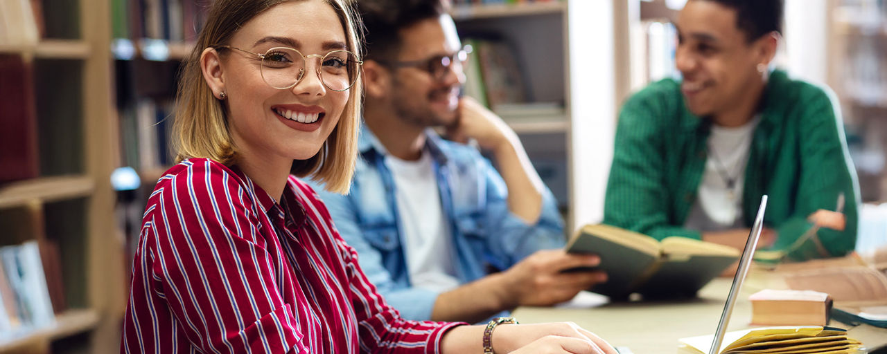  Drei Studierende lernen in der Bibliothek, eine Studentin sitzt vor ihrem Laptop und schaut in die Kamera.
