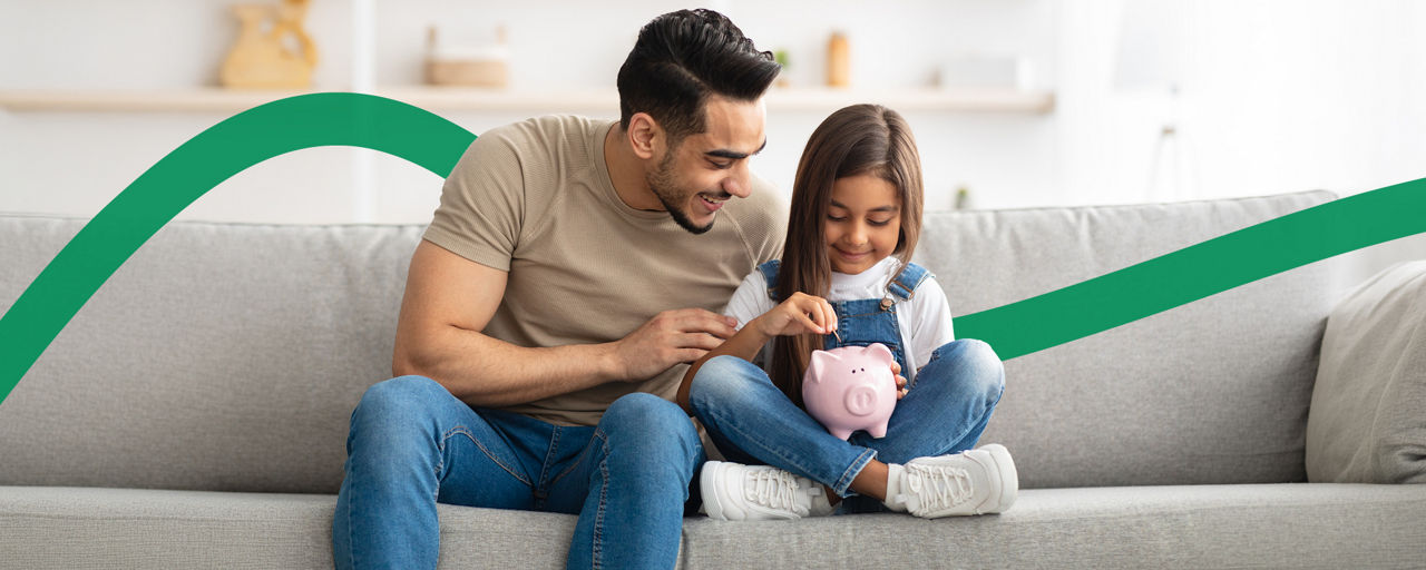 Ein Mann sitzt mit seiner Tochter auf dem Sofa und wirft Münzen in ein Sparschwein.