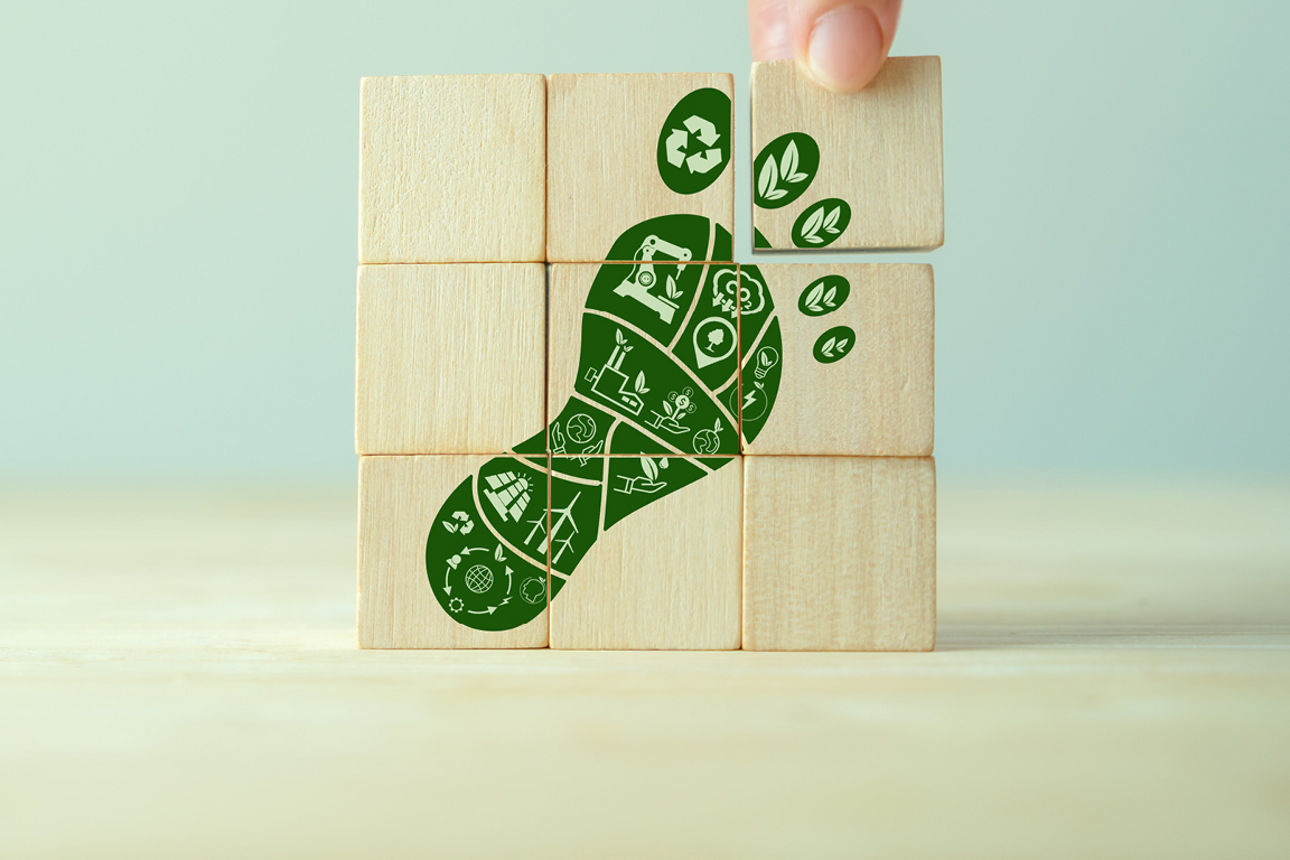 Zu sehen sind mehrere aufeinander gestapelte Holzwürfel, wo ein grüner Fußabdruck mit Umweltmotiven abgebildet ist.