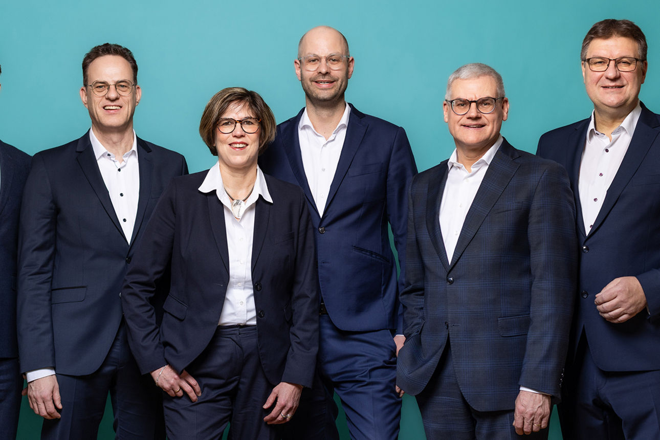 Der sechsköpfige Vorstand der neu entstandenen VR RegionalBank nebeneinanderstehend. Der Hintergrund ist petrolfarben.
