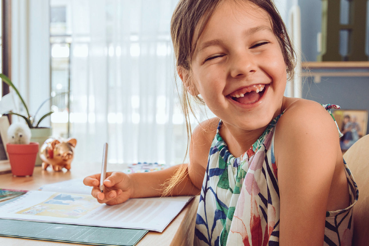 Ein junge Mädchen sitzt an einem Tisch und zeichnet etwas, sie sitzt halb umgedreht auf einem Holzstuhl und lächelt.