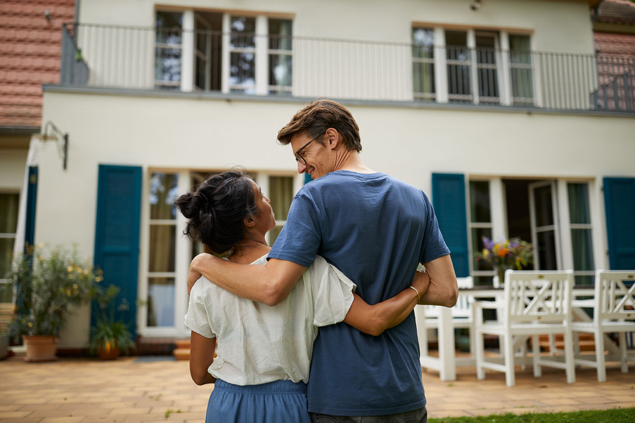Ein Paar steht Arm in Arm vor einem hellen Einfamilienhaus mit blauen Fensterläden, von hinten gesehen