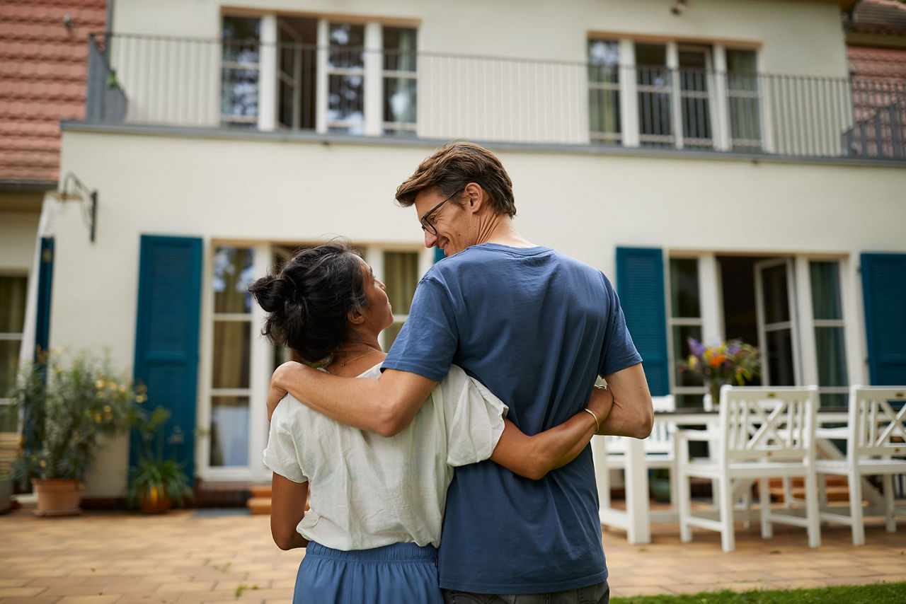 Zwei Personen stehen mit dem Rücken zur Kamera und haben sich umarmt. Sie blicken auf ein zweistöckiges Wohnhaus mit Balkon, großen Fenstern und einer Terrasse mit Tisch und Stühlen.