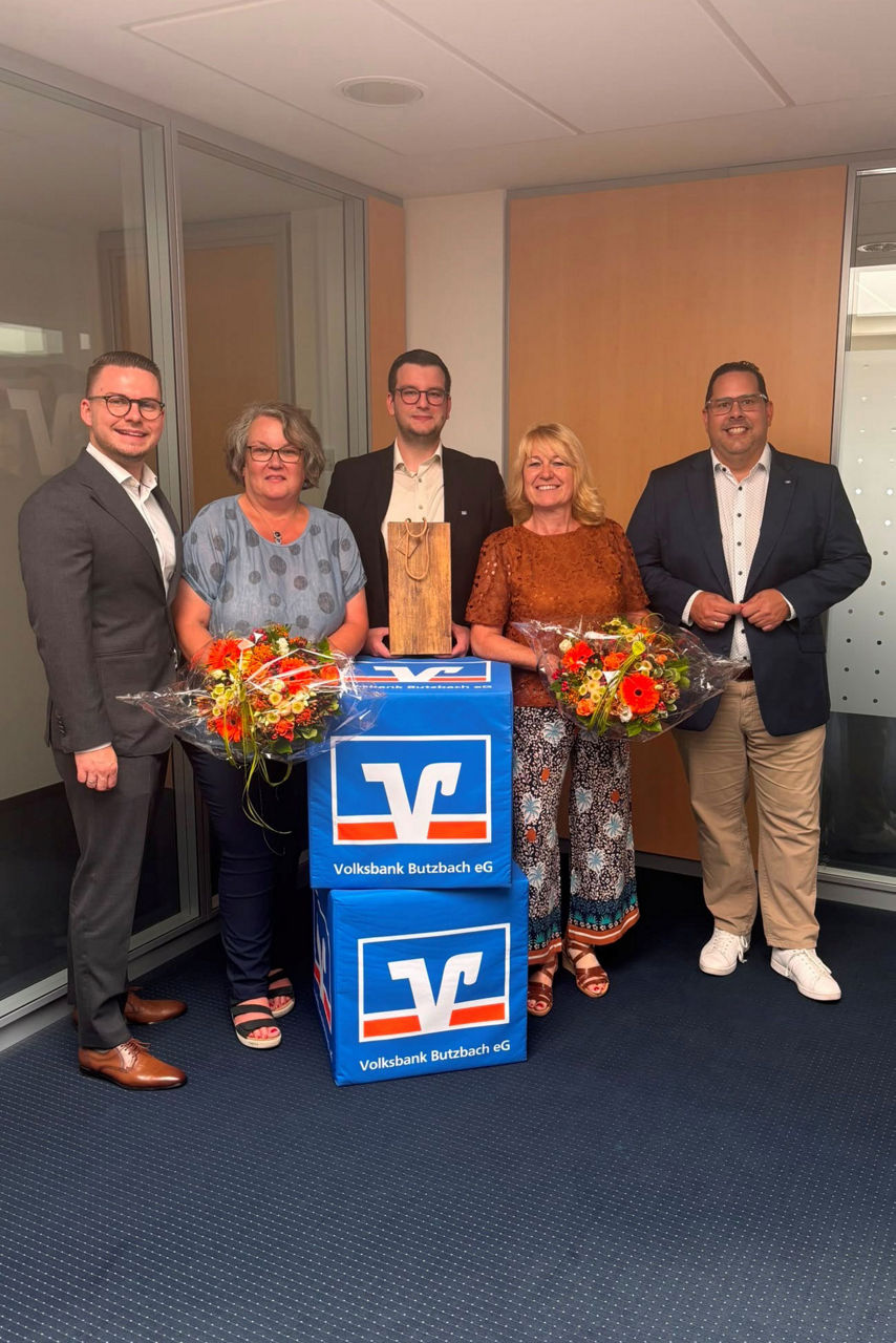 Zwei Mitarbeiterinnen der Volksbank Butzbach eG mit Blumensträußen zwischen drei Männern in Anzügen