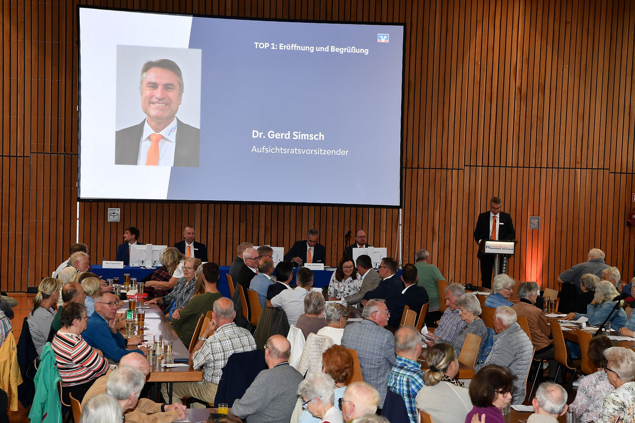 Gemeindeversammlung in einem Raum, Notizen und Getränke auf den Tischen sichtbar, während eine Präsentation stattfindet.