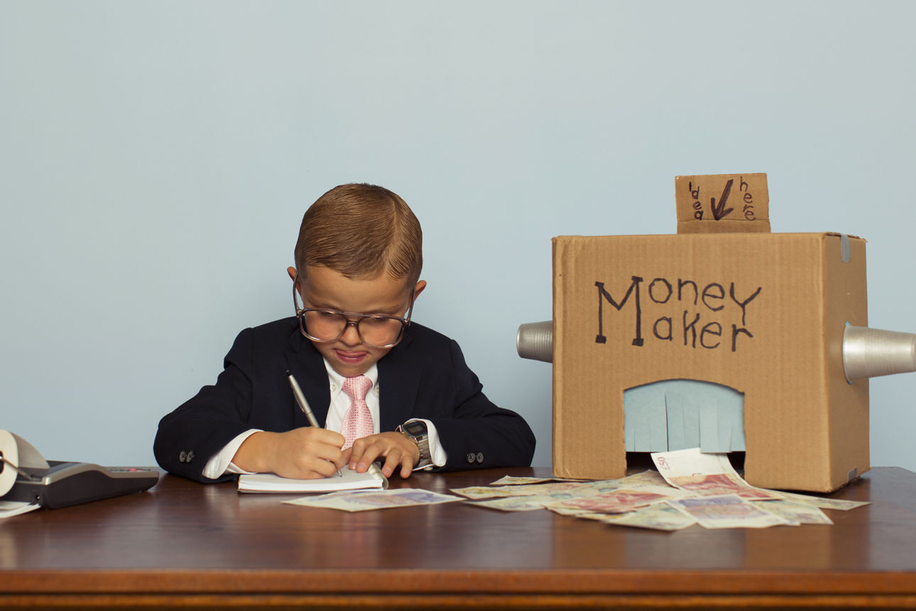 Ein Kind im Anzug an einem Schreibtisch. Auf einer Papp-Spardose steht "Money Maker"
