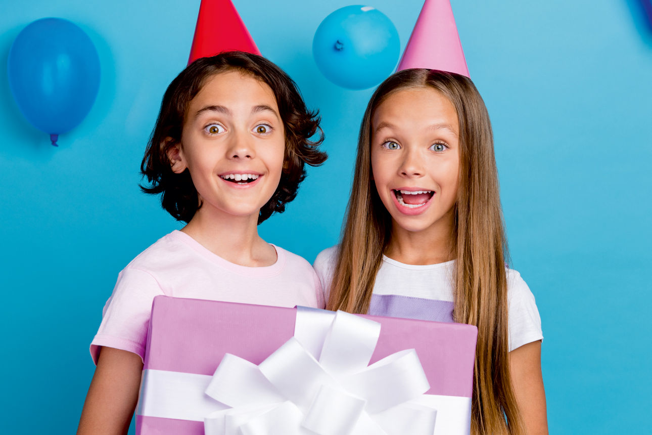 Zwei Kinder haben Partyhüte auf und stehen vor einer blauen Wand. Sie haben ein Geschenk mit Schleife in der Hand.