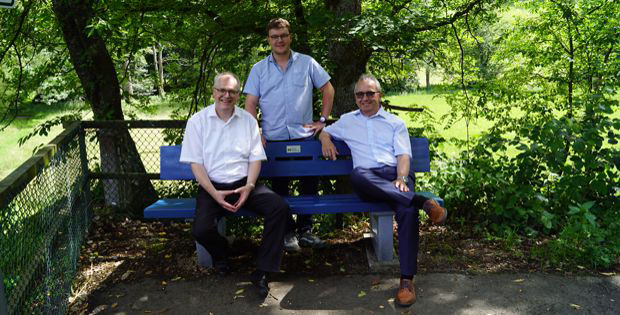 Drei Männer sitzen auf einer blauen Bank in einem Park, sie tragen alle helle Hemden und dunkle Hosen.