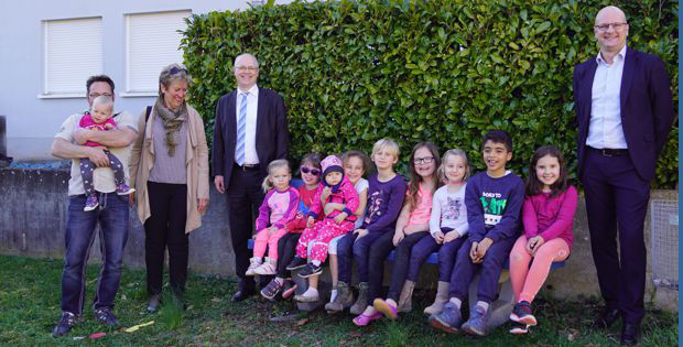 Eine Gruppe von Kindern, einige sitzend auf einer Bank, andere stehend, versammelt sich draußen mit mehreren Erwachsenen.