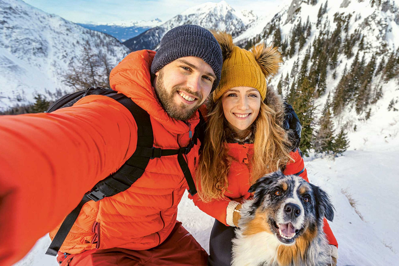 Ein Mann, eine Frau und ihr Hund machen bei winterlichem Wetter in den Bergen ein fröhliches Selfie.