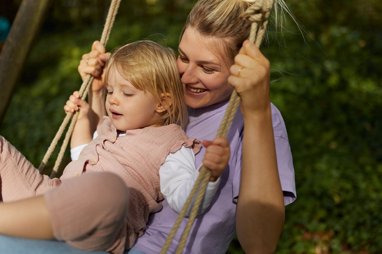 Erwachsene Person hält ein Kind auf einer Schaukel im Freien, umgeben von grünem Hintergrund.
