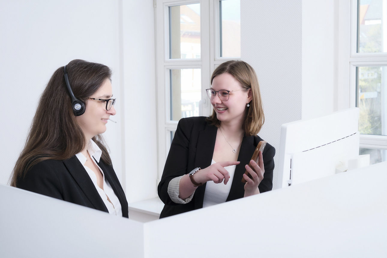 Zwei junge Frauen - eine mit Headset - stehen an einem PC 