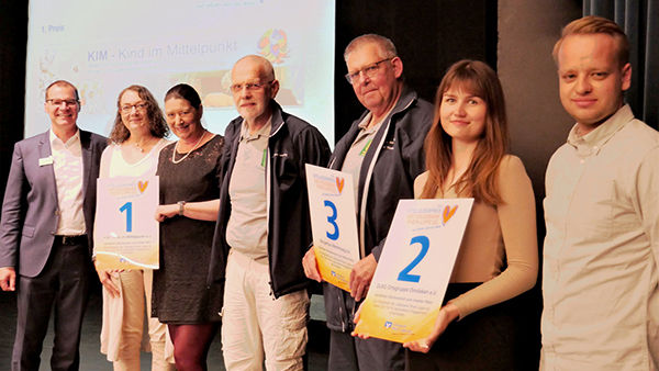 Eine Gruppe von sechs Personen steht in einer Reihe und hält Zertifikate mit den Nummern 1, 2 und 3 hoch.
