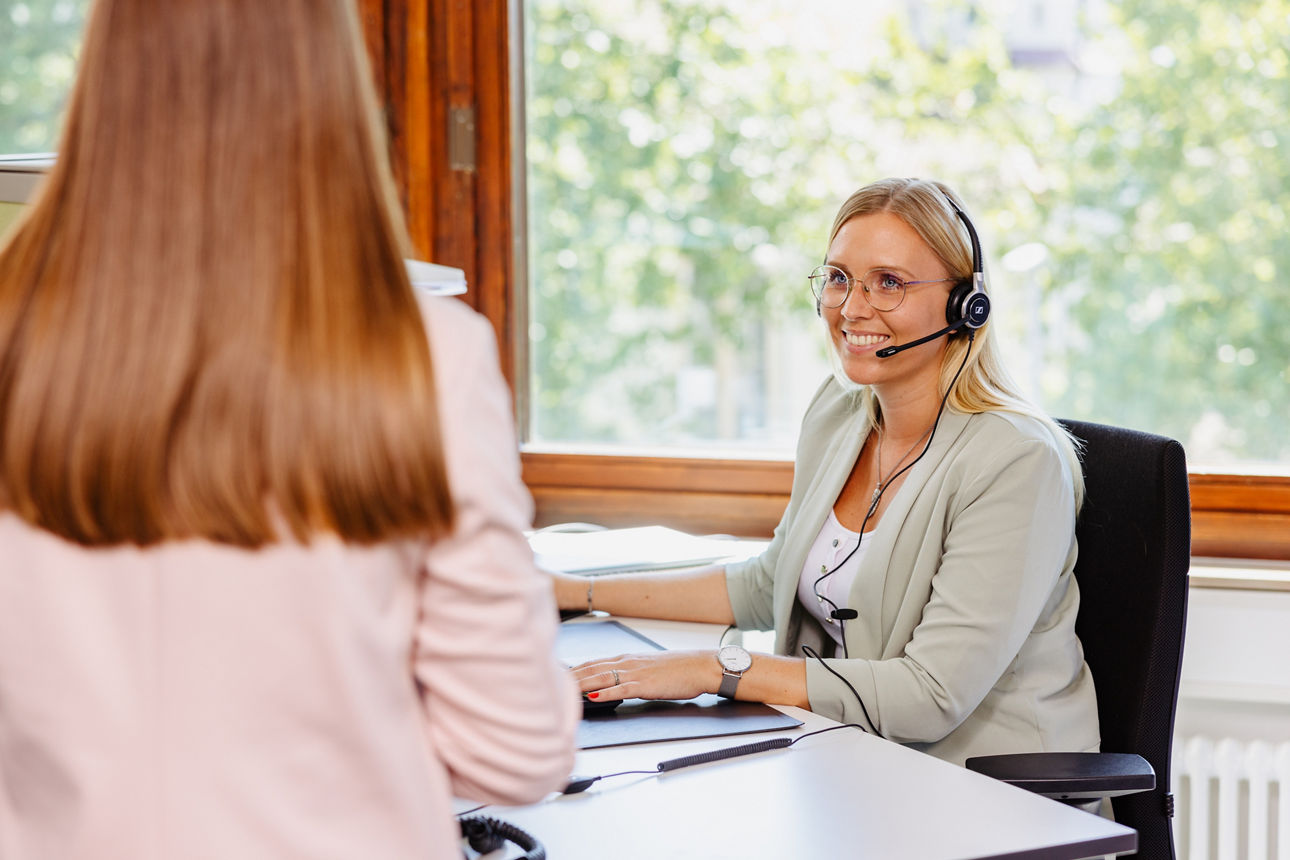 Lächelnde Frau mit Headset sitzt an einem Schreibtisch.