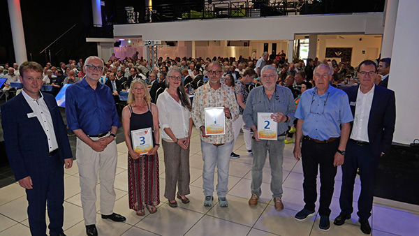 Eine Gruppe von acht Personen steht in einer Reihe und hält Zertifikate mit den Nummern 1, 2 und 3 hoch.