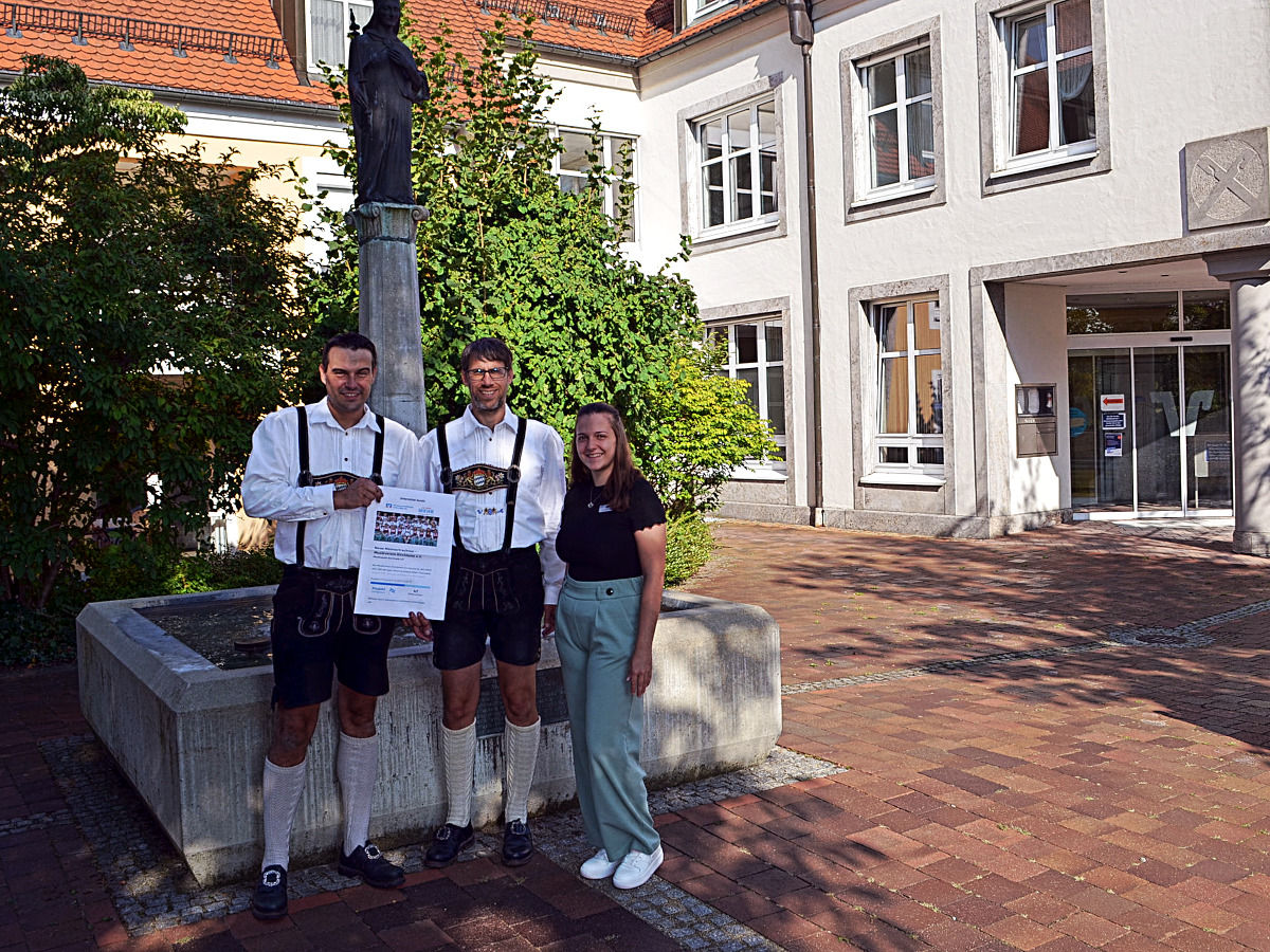 Zwei Ansprechpartner von der Musikkapelle stehen nebeneinander mit einer Mitarbeiterin unserer Bank vor einem Brunnen