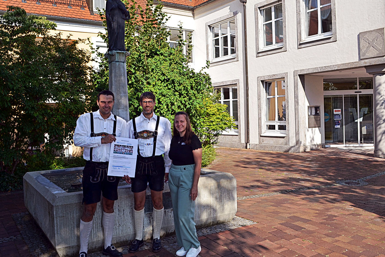 Zwei Ansprechpartner von der Musikkapelle stehen nebeneinander mit einer Mitarbeiterin unserer Bank vor einem Brunnen