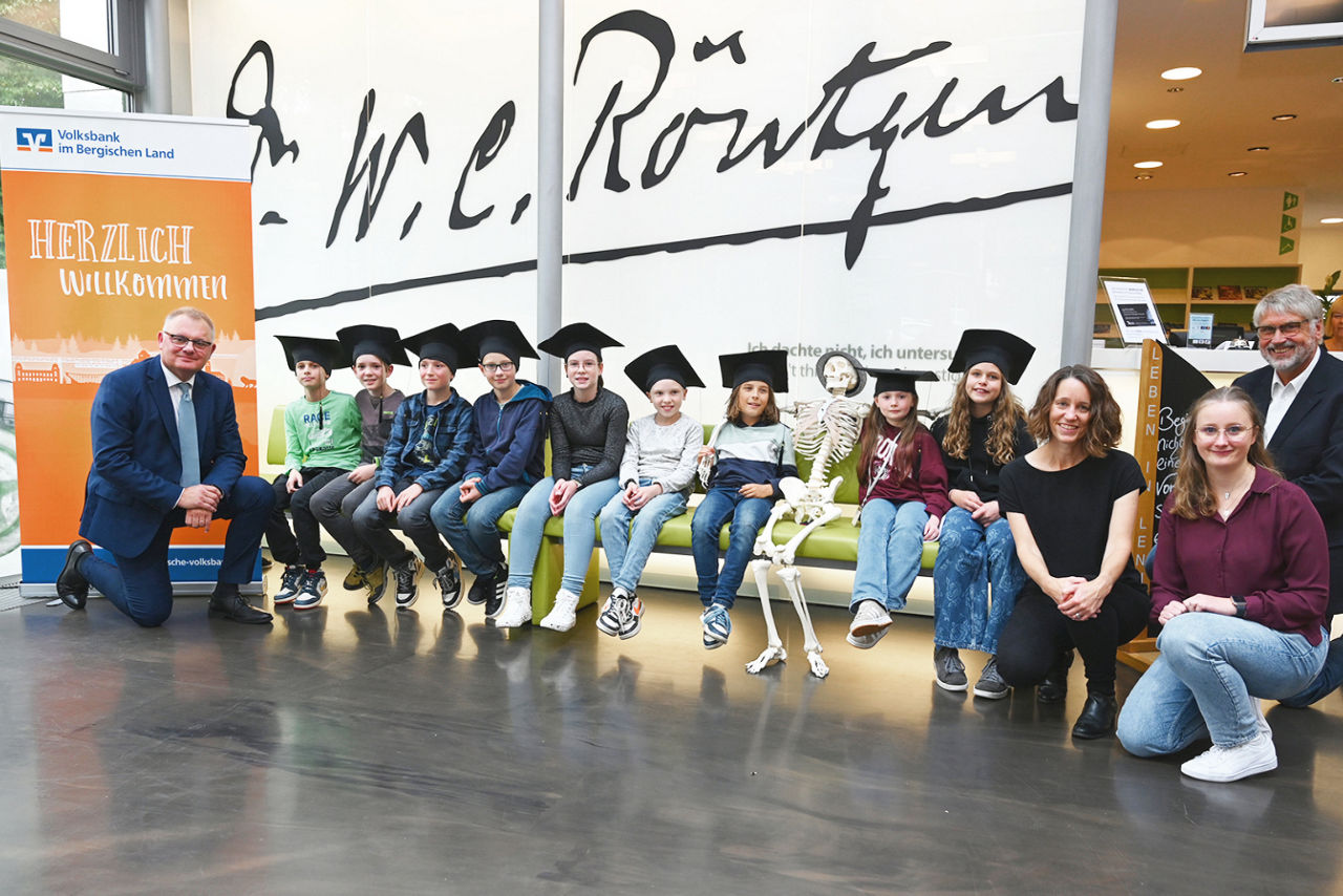Kinder mit Bachelorhüten sitzten mit Vorstand auf Bank im Mueseum