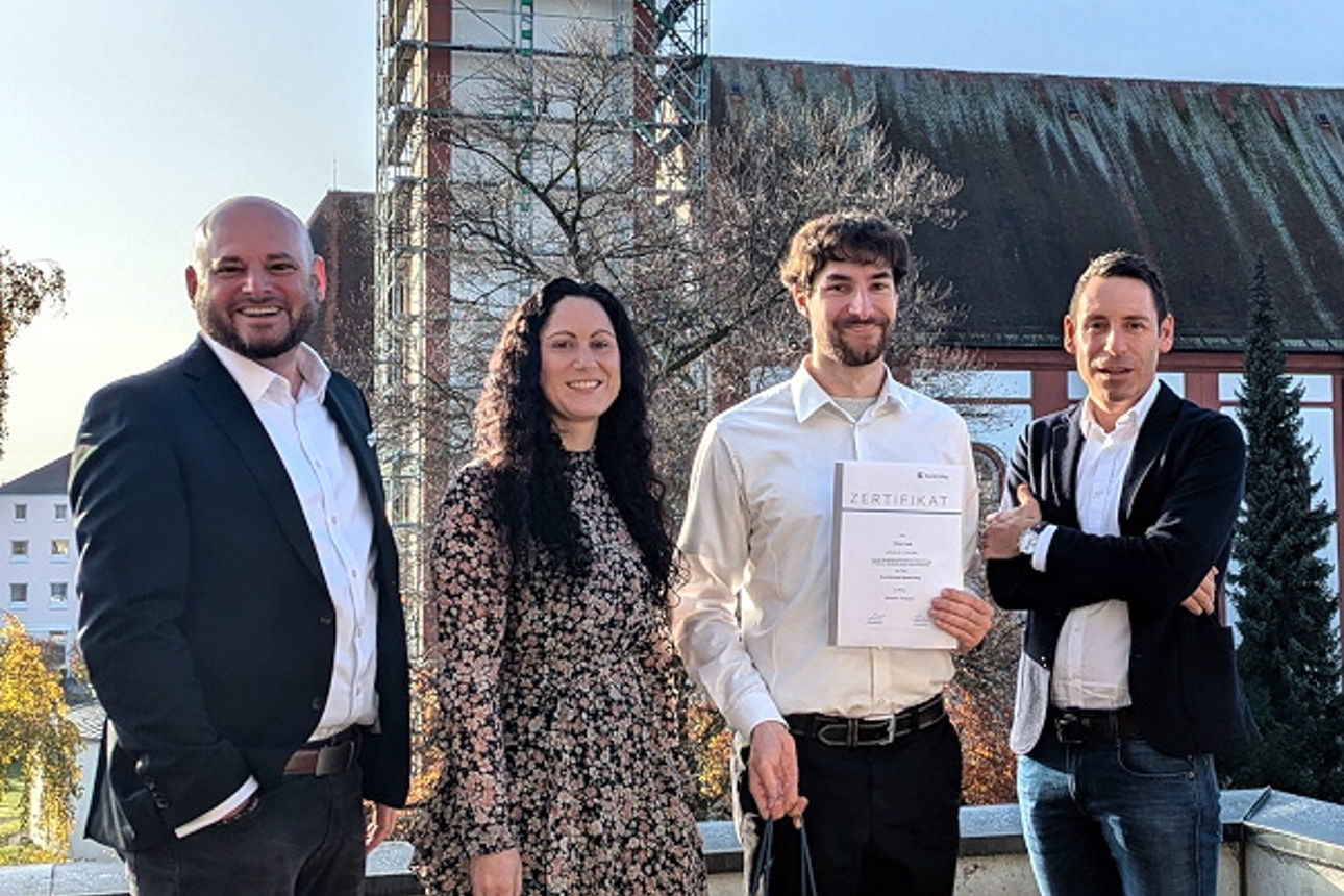 Zwei Mitarbeiter der Bank stehen mit Urkunde neben dem Vorstand vor einem Kirchturm