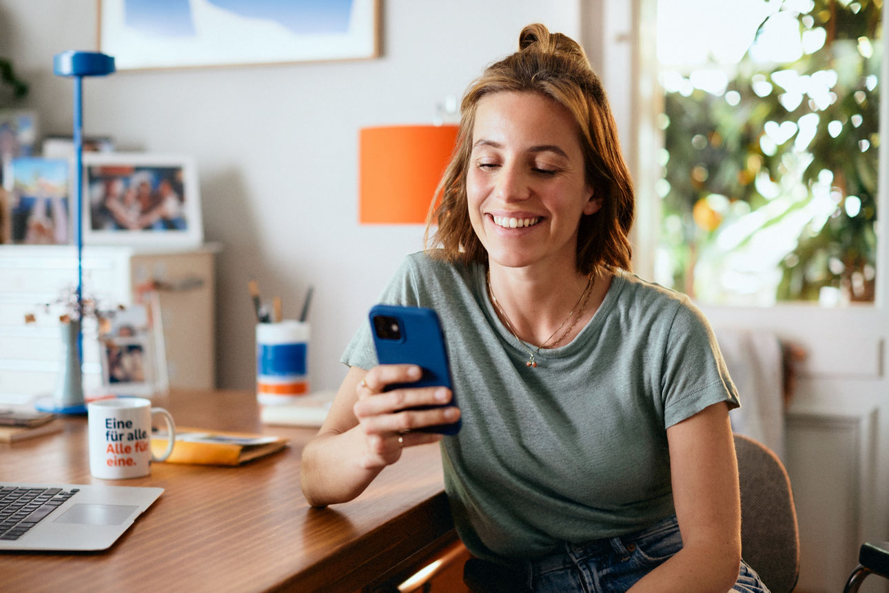 Frau mit Smartphone in der Hand an einem Schreibtisch sitzend.