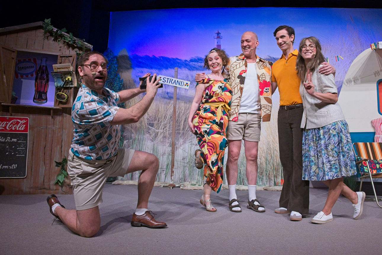 Eine Gruppe von Komödien verkleidet in Strandkleidern stehen Arm in Arm und lassen sich von dem Kamermann fotografieren