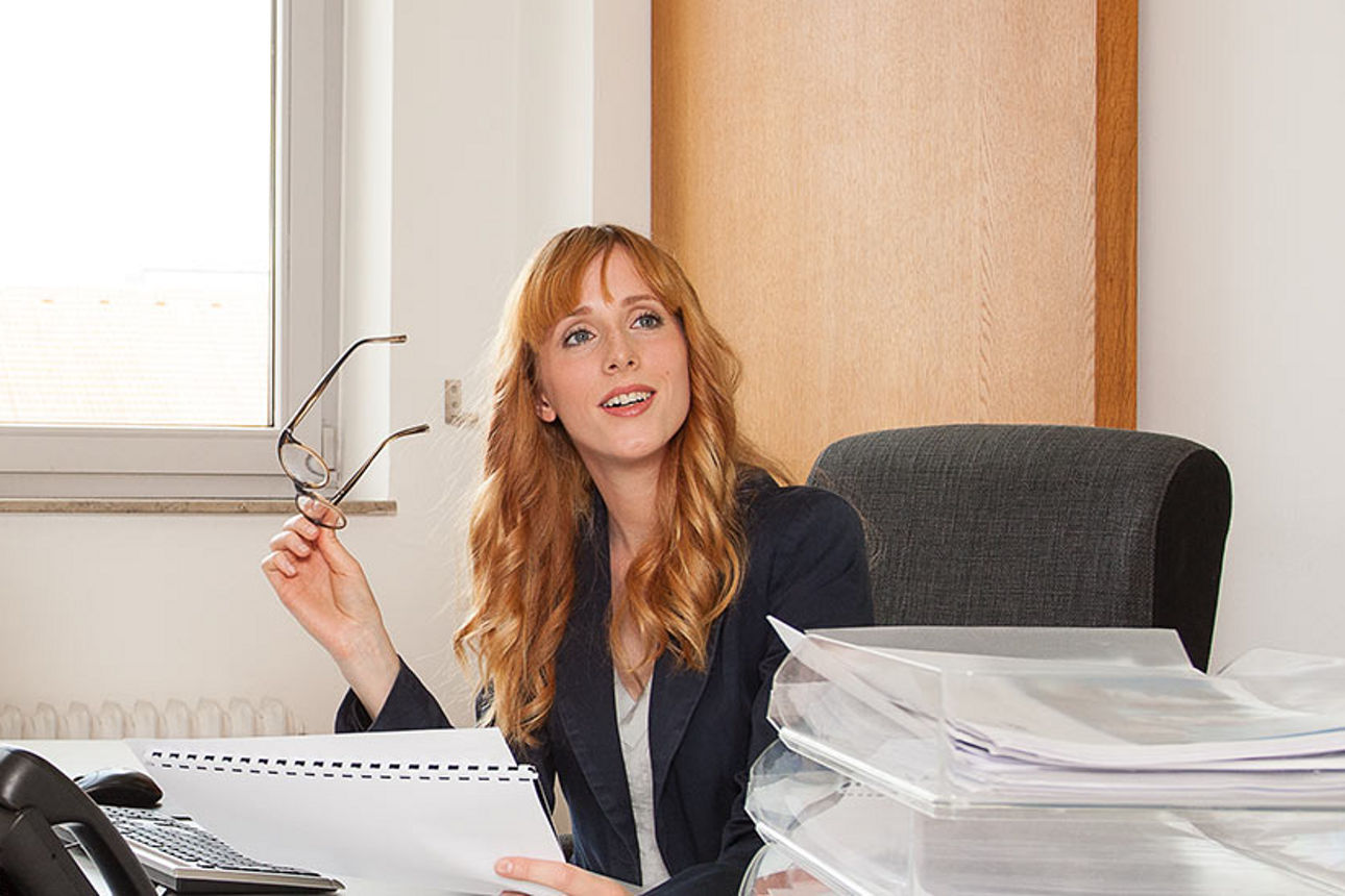 Eine Frau schaut von Ihrem Schreibtisch auf während sie ihre Brille in der Hand hält