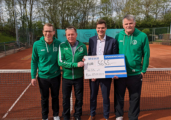 Vier Männer stehen auf einem Tennisplatz, davon drei im Sportanzug und bekommen einen Scheck überreicht