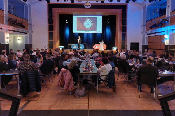 Publikum sitzt  in einem Saal und schaut auf eine Bühne