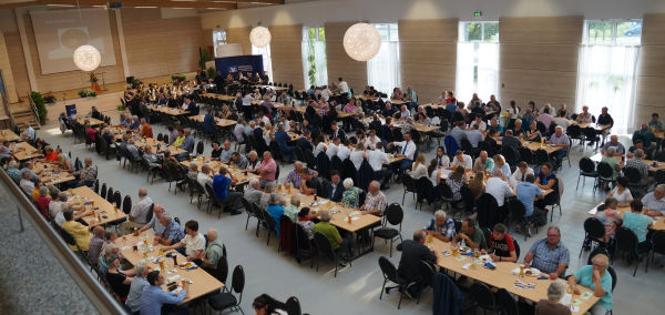 Eine große Gruppe von Menschen, die in einem geräumigen Raum an Tischen sitzt und sich unterhält und beschäftigt.