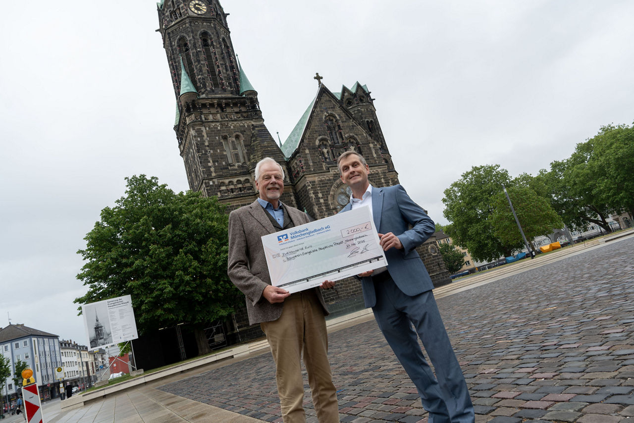 Zwei Männer halten einen Spendenscheck vor der Kirche. Die Volksbank unterstützt die Sanierung der Turmspitze mit 2.000 Euro.