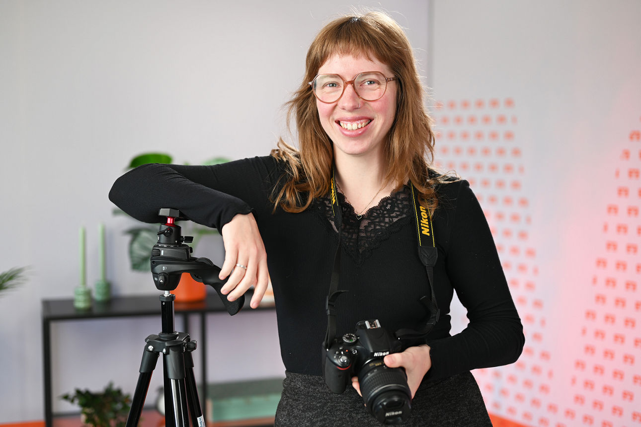 Alicia steht im Videostudio, lehnt sich am Stativ an und hat eine Kamera in der Hand