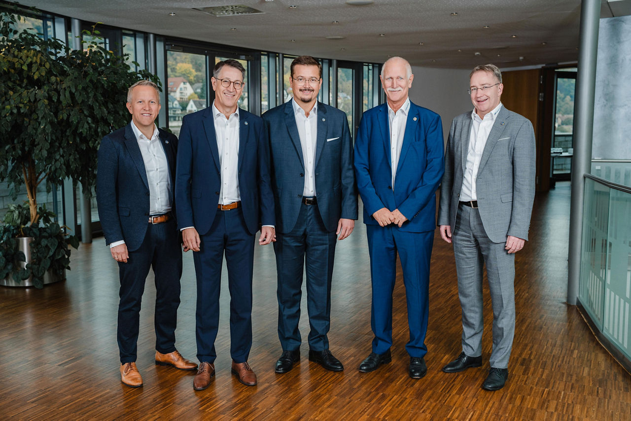 Fünf Männer stehen nebeneinander und lächeln in die Kamera. Sie tragen Anzüge in verschiedene Blautöne und Grau.