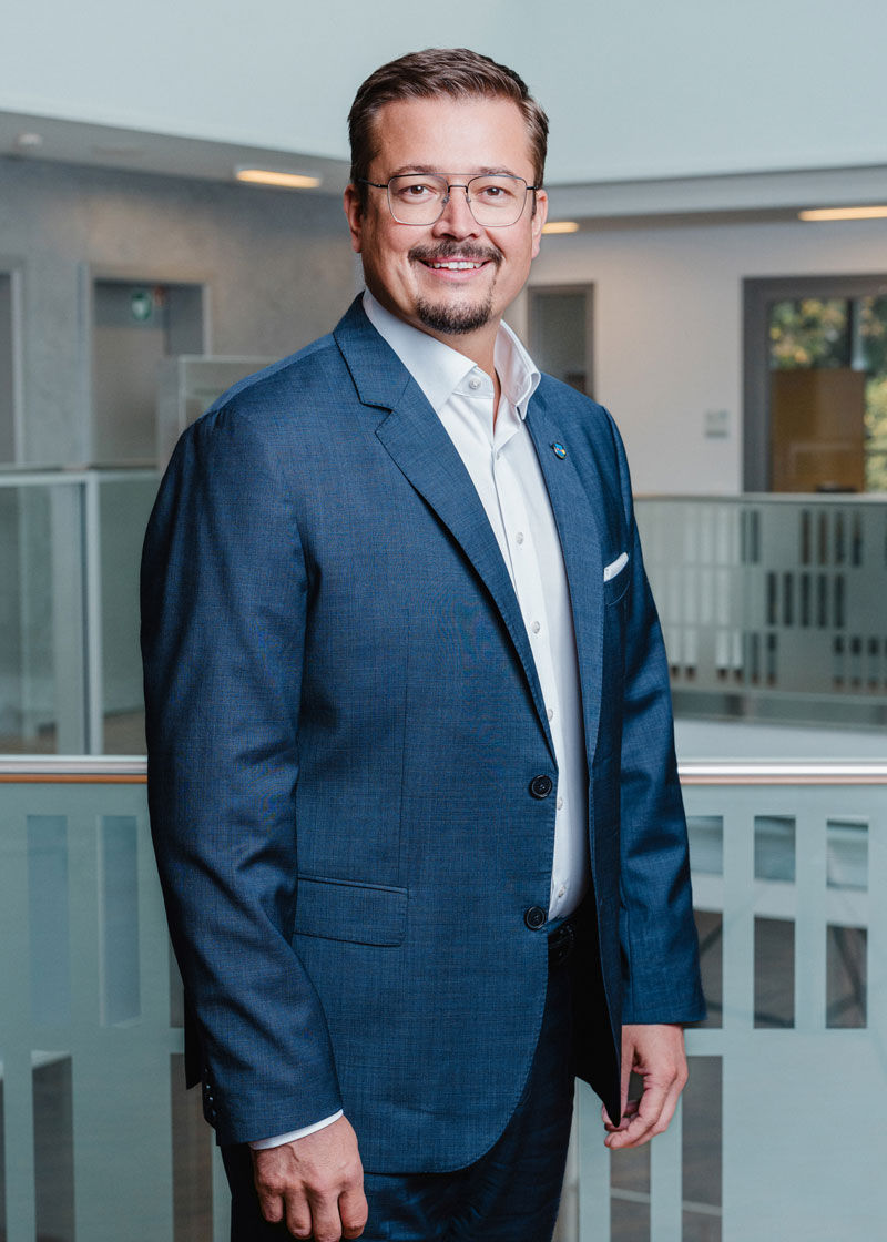 Ein Mann steht im dunklen Anzug vor einer Glaswand und lächelt in die Kamera.