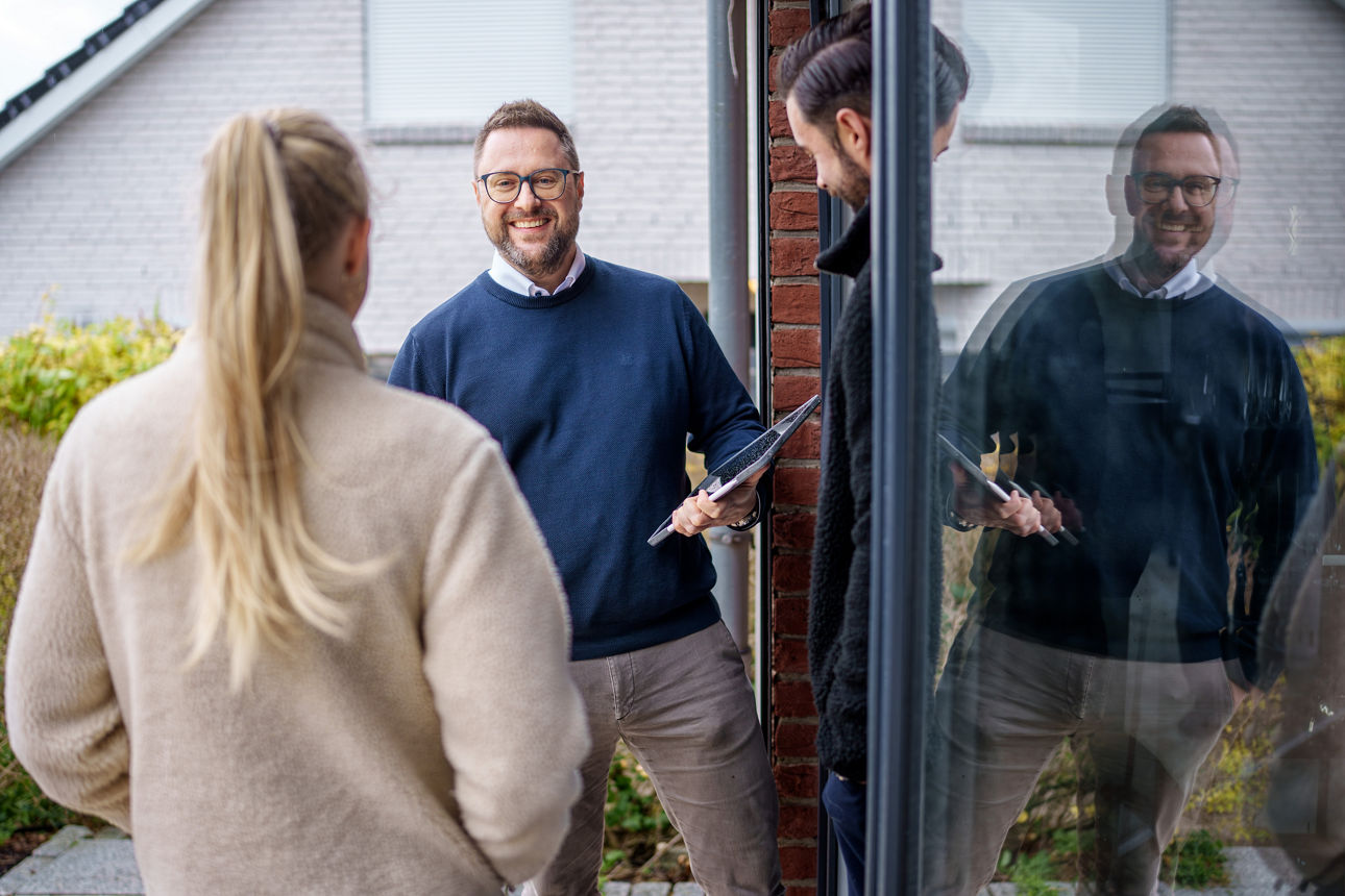Drei Personen stehen vor einem Haus, eine Frau mit hellem Mantel und Pferdeschwanz von hinten, zwei Männer, einer hält ein Tablet.
