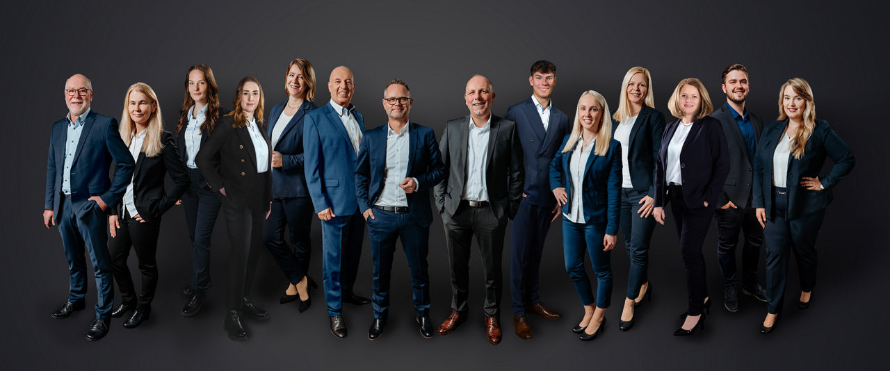 Das Team der Immobilienexperten steht vor einem schwarzen Hintergrund. Es besteht aus 14 Personen, die Business Kleidung in blau und schwarz tragen.
