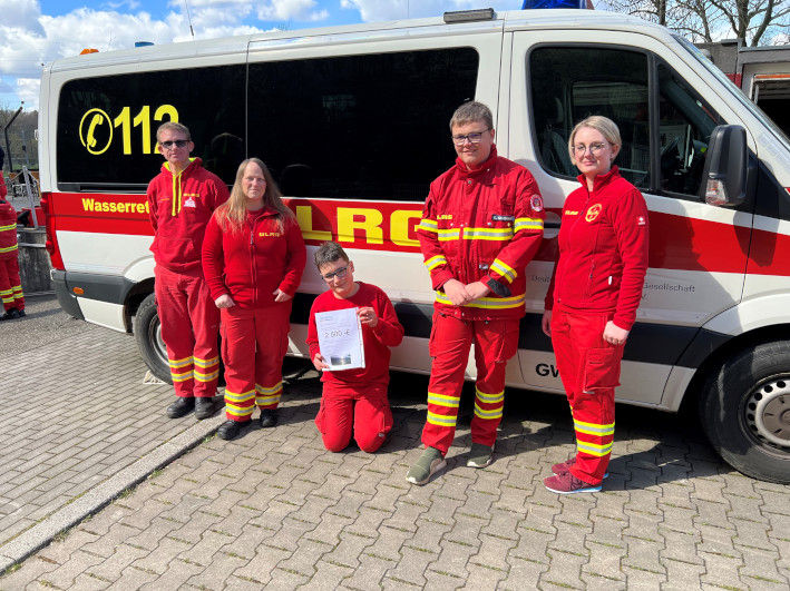 Mitglieder des DLRG in Uniform vor ihren Einsatzwagen mit einem Scheck in der Hand