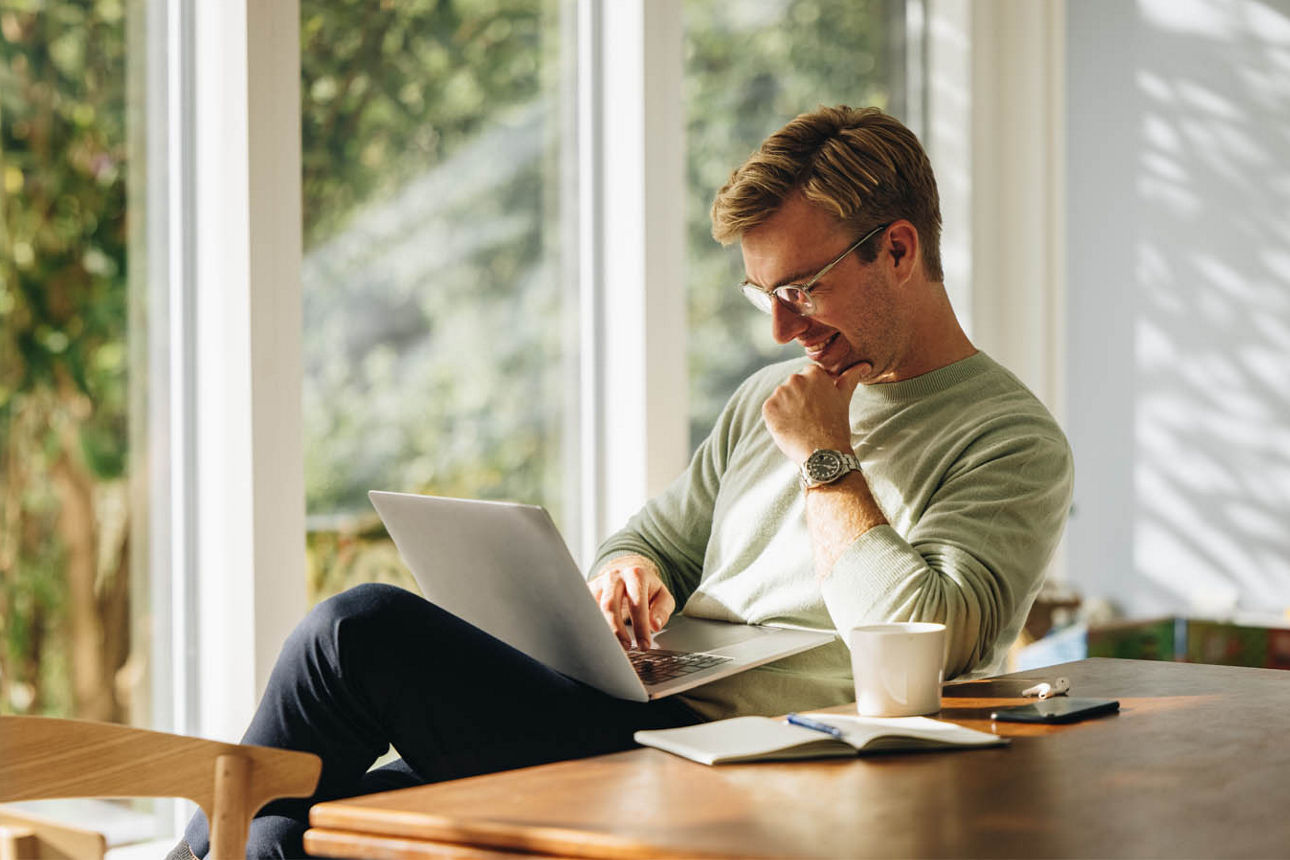 Junger Mann sitzt lächelnd vor seinem Laptop